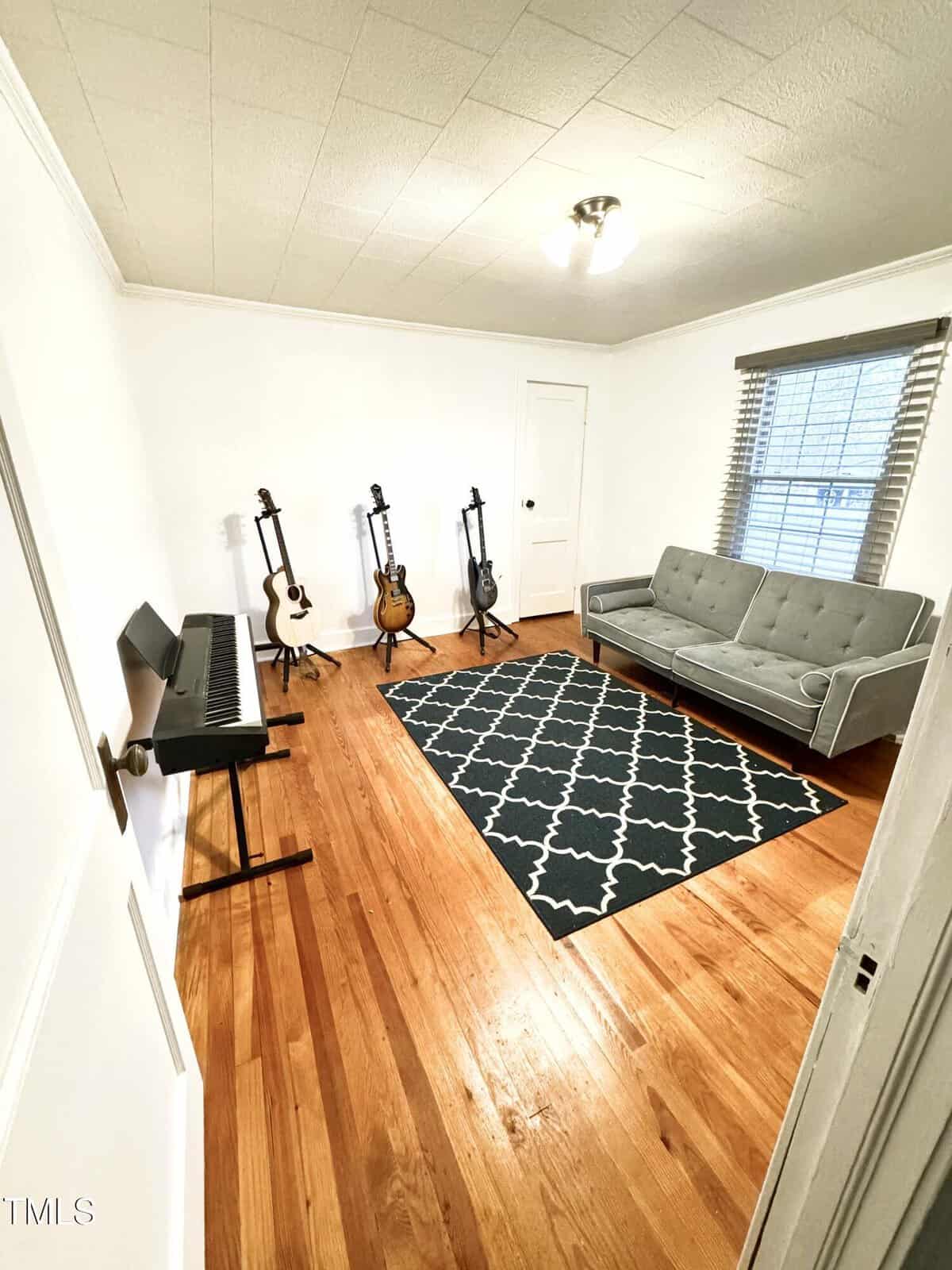 A room featuring a keyboard on a stand, four guitars on stands, a gray sofa, and a patterned dark rug on a wooden floor. The walls are white, and a window with blinds is on the right side.