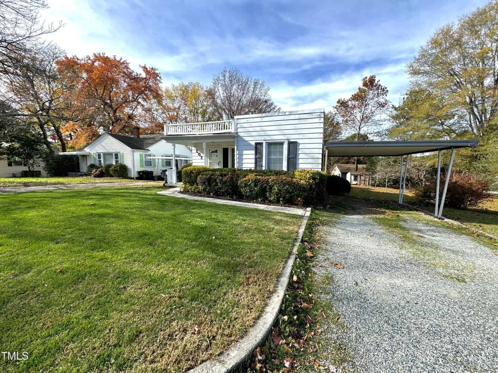 A residential house with a white exterior, surrounded by a neat lawn and autumn trees. There is a gravel driveway leading to a metal carport on the right side, and a small porch at the front of the house. The sky is partly cloudy.