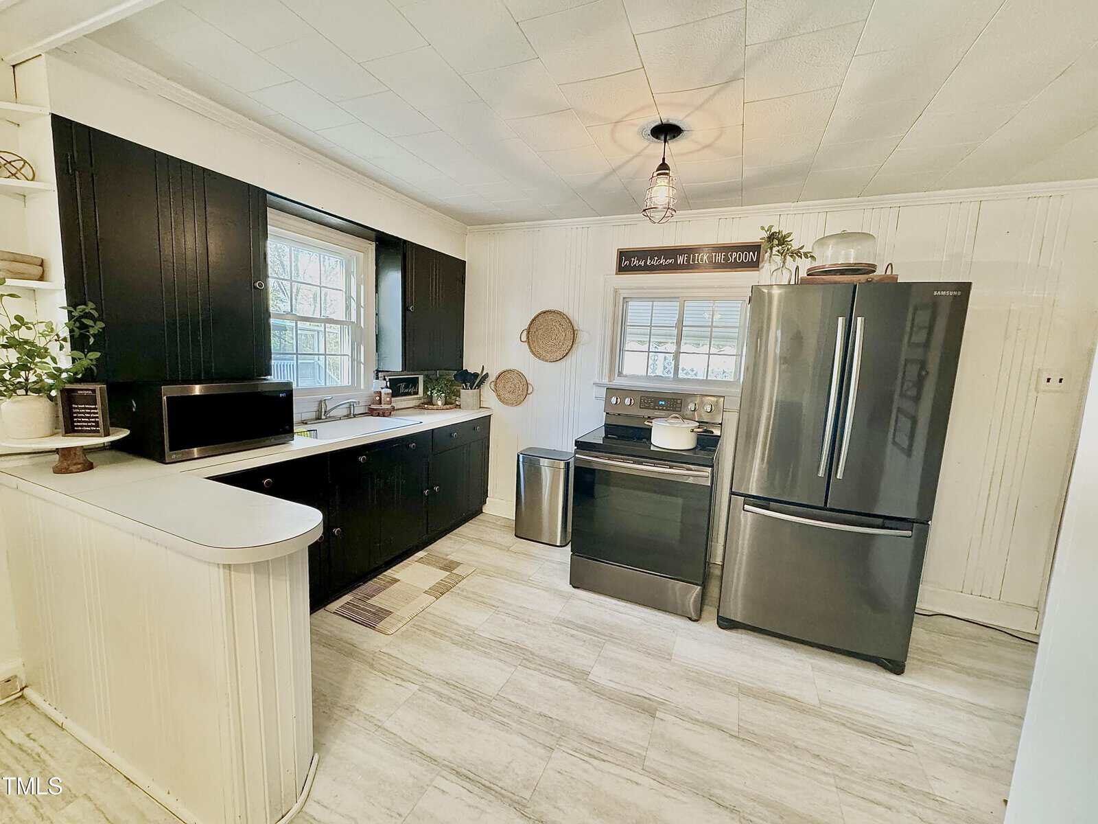 Kitchen with white and black decor. Features a stainless steel refrigerator and oven, black cabinets, and a white countertop. Two windows provide natural light. A light fixture hangs from the ceiling, and various plants and baskets decorate the space.