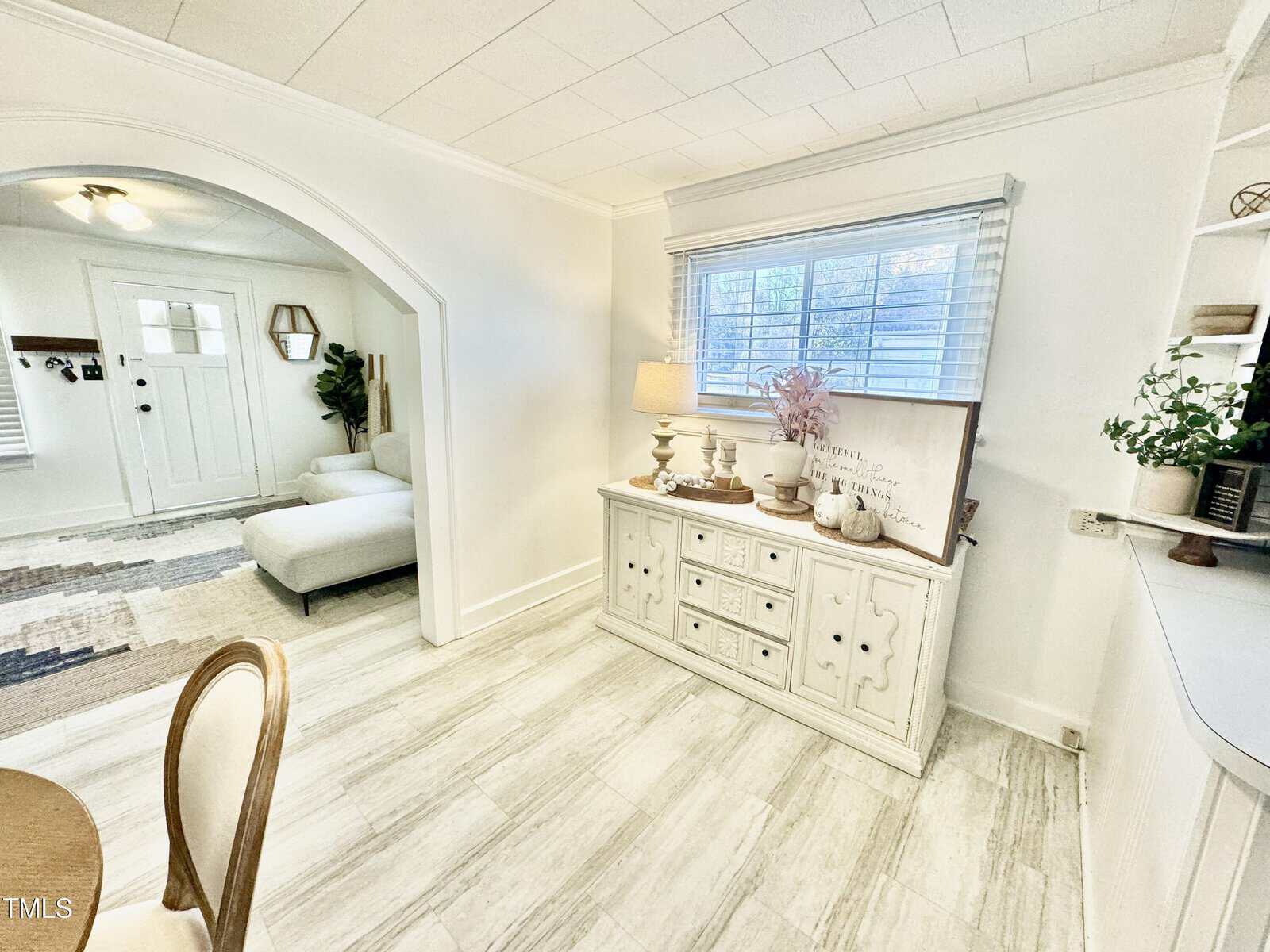 A bright room with white walls and light wood flooring features a wooden sideboard with decorative items, including a lamp and a sign. A window with blinds is above the sideboard. An archway leads to a seating area with a grey sofa.