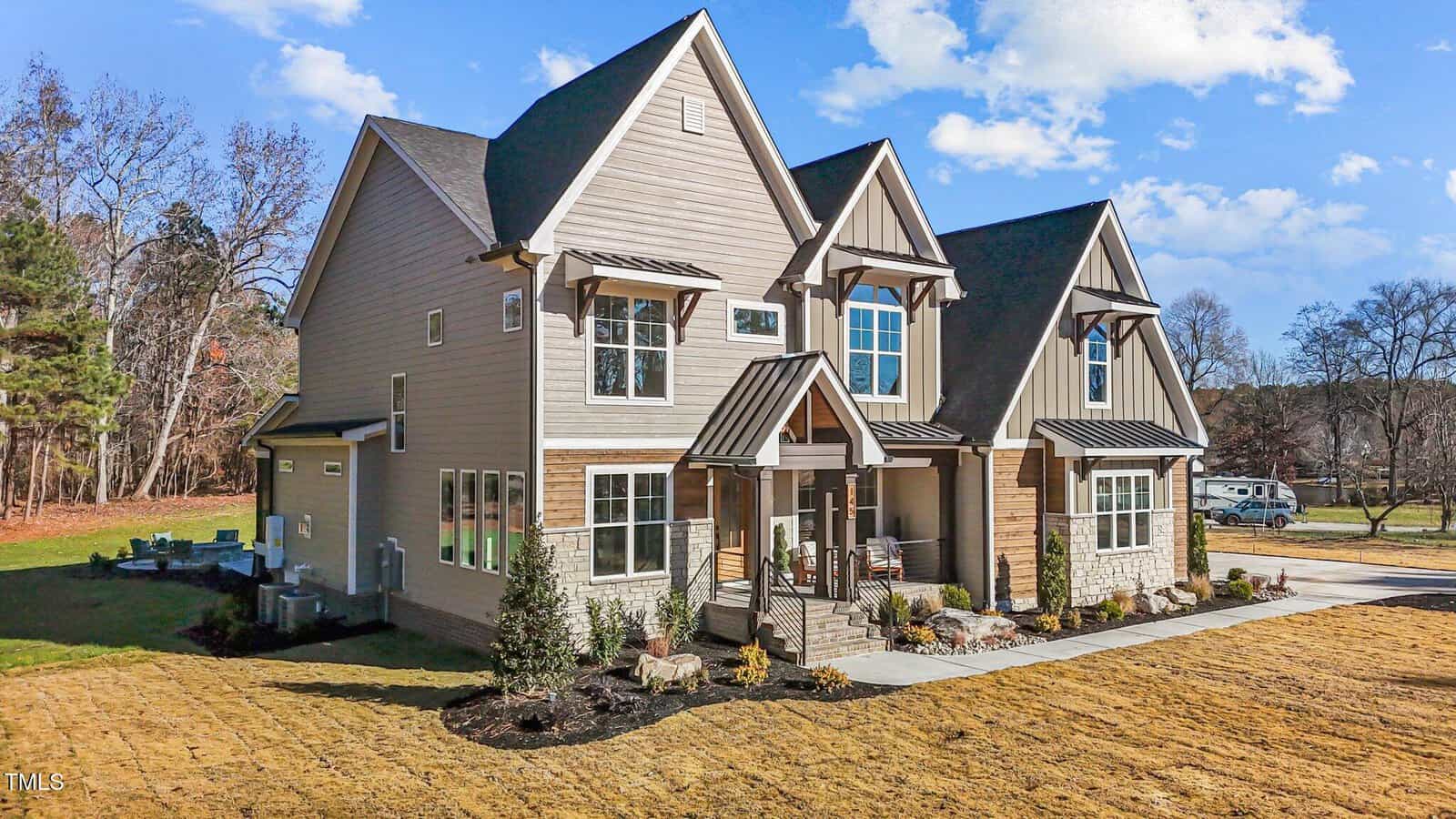 Two-story modern house with multiple peaked roofs, large windows, and a covered front porch. The home is surrounded by landscaped shrubs and grass, with a driveway and a wooded area in the background under a blue sky.