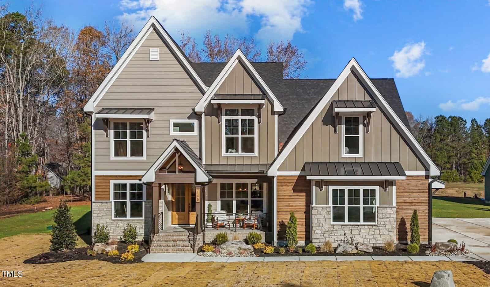 Two-story modern house with beige and brown siding, stone accents, three prominent front gables, large windows, covered porch with columns, small landscaped front yard, and driveway on the right at 145 Wiggins Road in Youngsville. Trees and blue sky in the background.