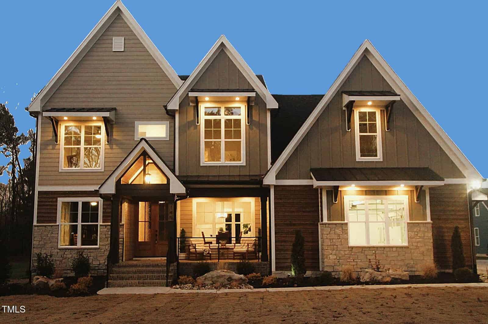 A two-story house with peaked roofs, large windows, stone and wood exterior, and a covered entrance. Outdoor lights are on, and the sky is clear and blue. Shrubs and rocks line the front walkway.