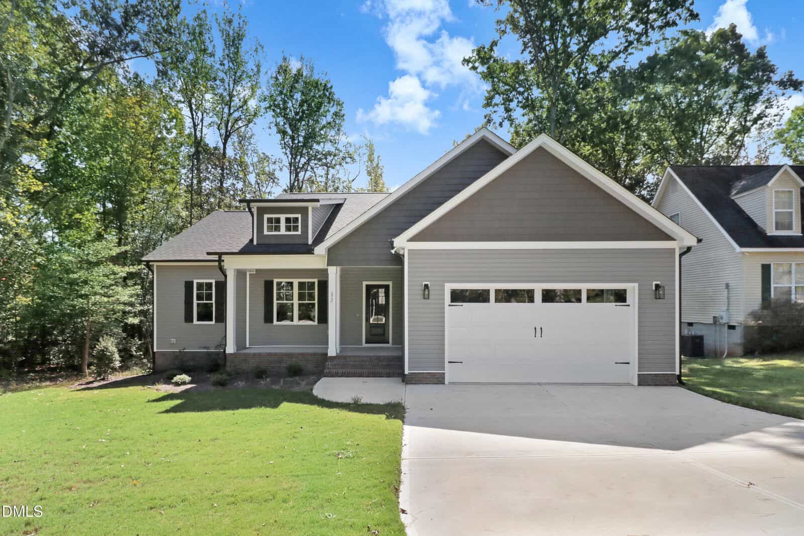 A gray, single-story house with white trim, a double garage, and a concrete driveway sits at 652 Shawnee Drive in Louisburg. The front yard features green grass and is surrounded by trees, while the sky above is blue with some clouds.