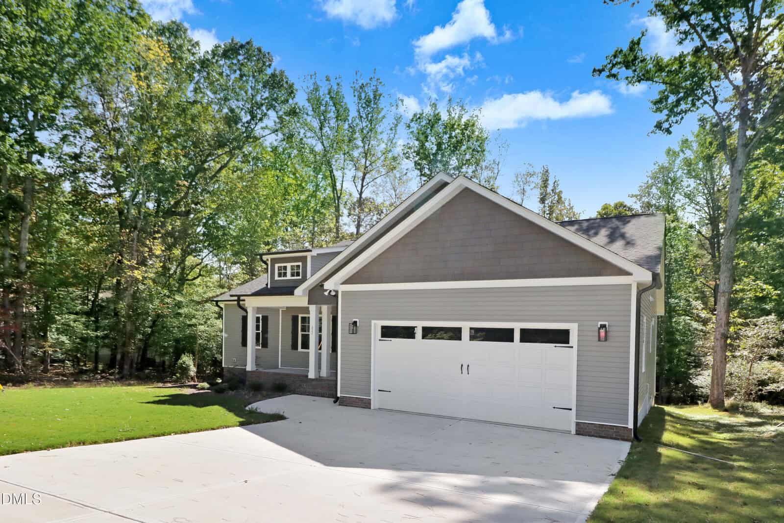 Single-story gray house with white trim, attached double garage, and front porch, set on a concrete driveway. Surrounded by trees and green grass, with a clear blue sky in the background.