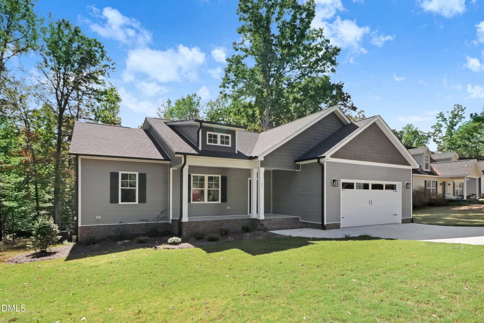 Single-story gray house with white trim, attached two-car garage, front porch, and a large lawn. Surrounded by trees under a blue sky with some clouds. Neighboring houses are partially visible in the background.