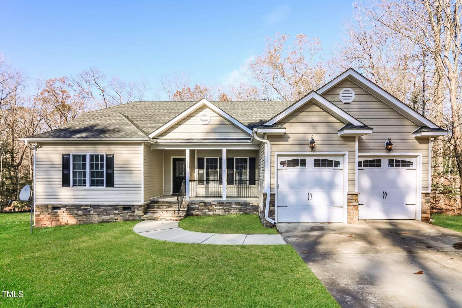 Single-story house with beige siding, stone accents, and a covered porch nestled at 186 Buckaroo Drive, Louisburg. There are two white garage doors and a curved walkway leading to the front steps, surrounded by a lawn and trees with sparse leaves.