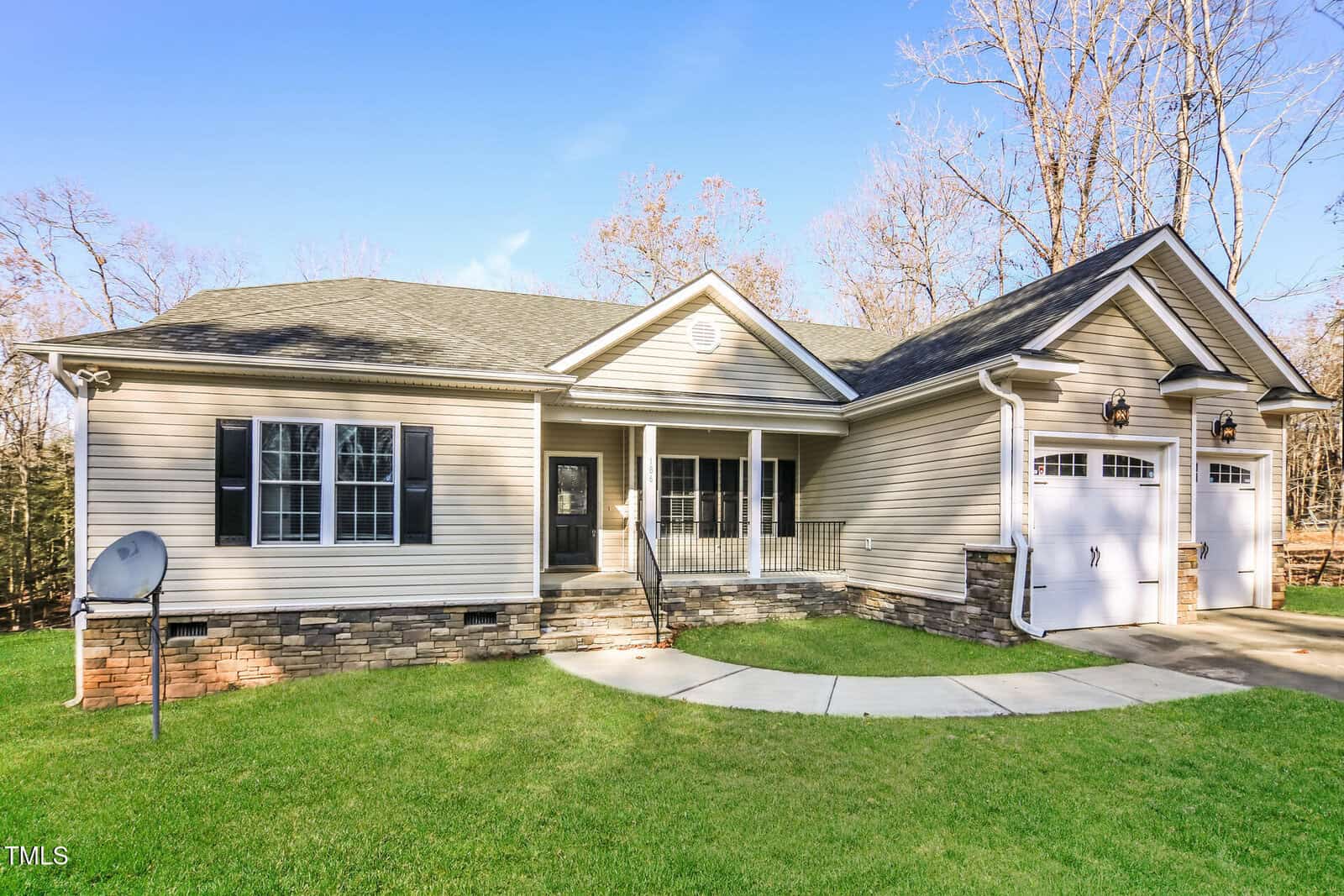 Single-story beige house with black shutters, a covered front porch, stone accents, double garage, and a curved sidewalk. The lawn is green, and there are bare trees in the background under a clear blue sky.