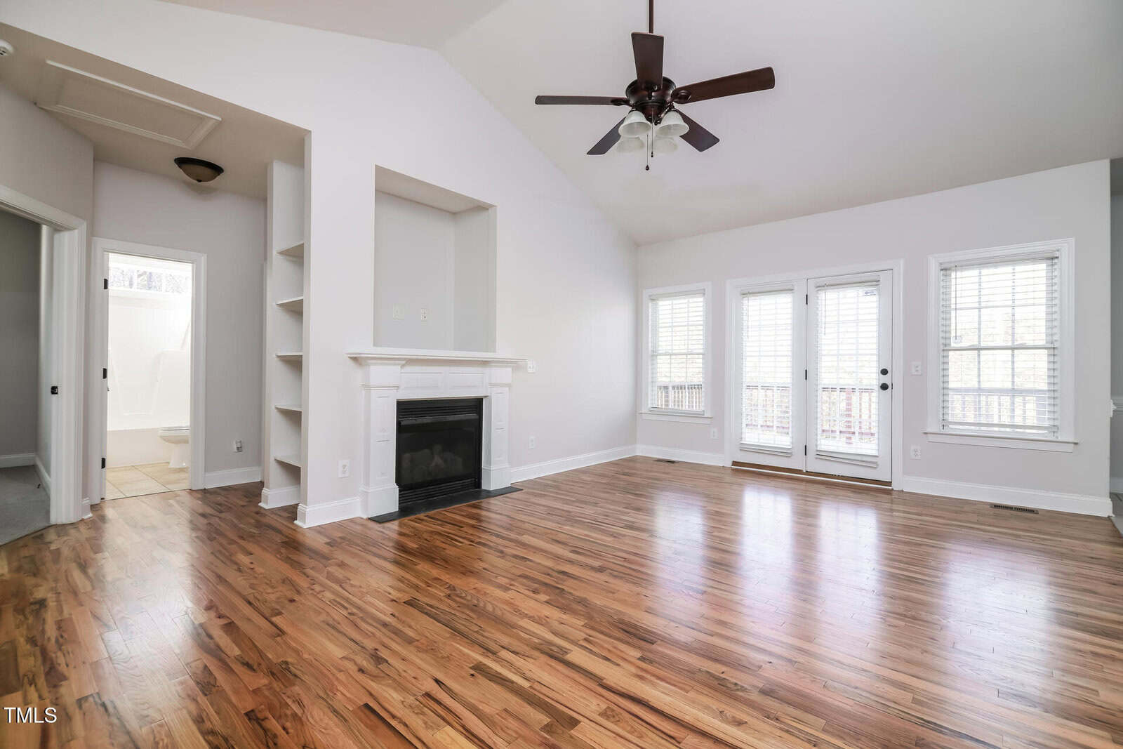 Bright living room with hardwood floors, a ceiling fan, fireplace, built-in shelves, and large windows with white blinds. French doors lead to a patio, and a bathroom is visible through an open door.