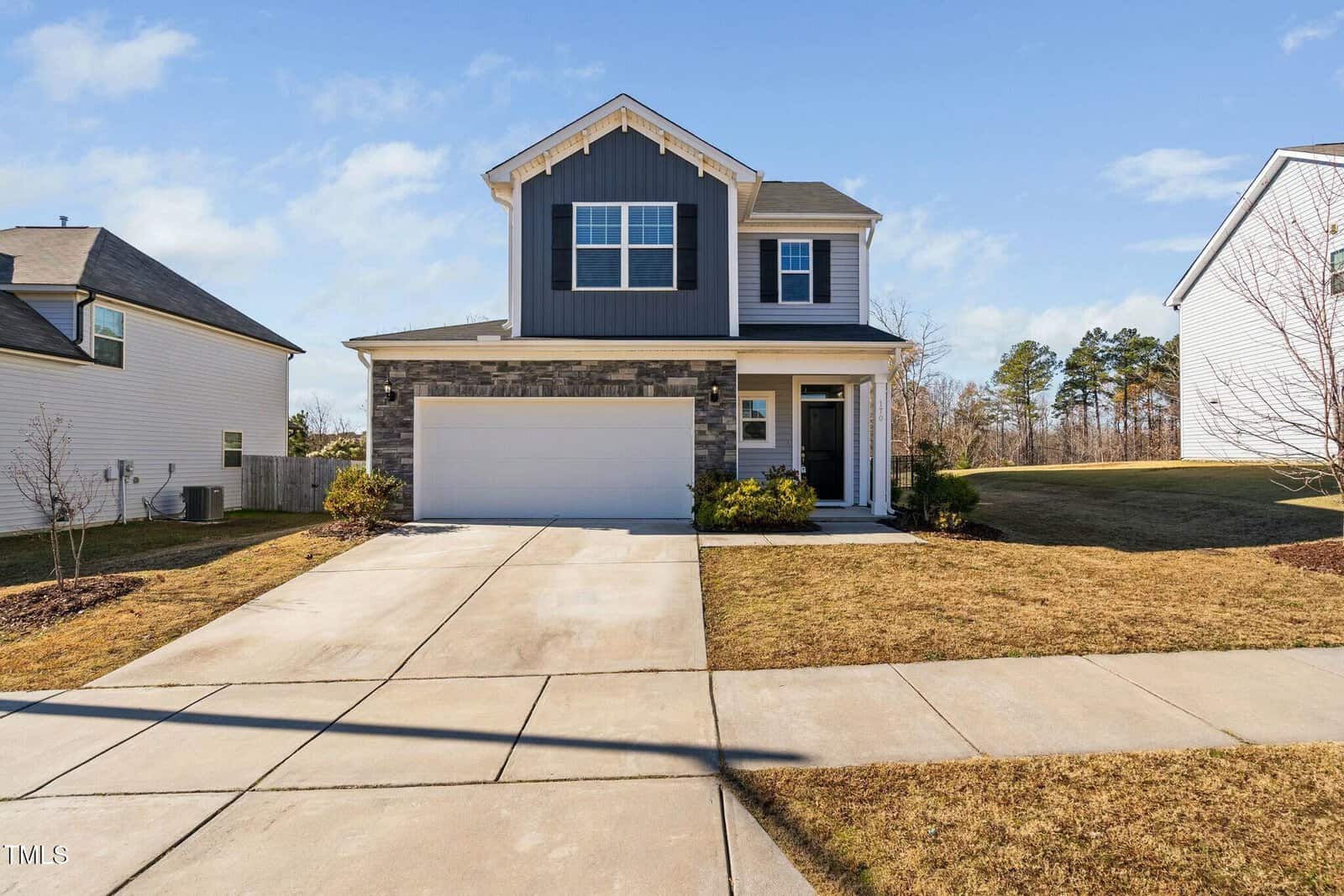 Two-story suburban house with gray siding and stone accents, white trim, and a two-car garage. The driveway and lawn are neatly maintained, and neighboring houses are visible on both sides. Blue sky with light clouds overhead.