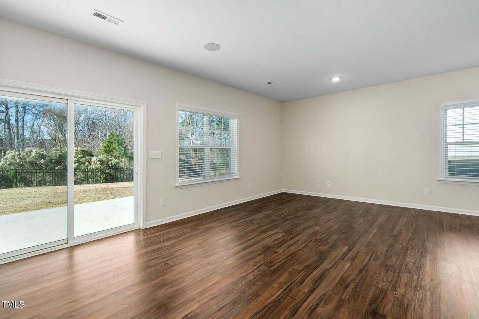 A spacious, empty room with light beige walls, wood flooring, large windows, and a sliding glass door opening to a patio and backyard with trees.