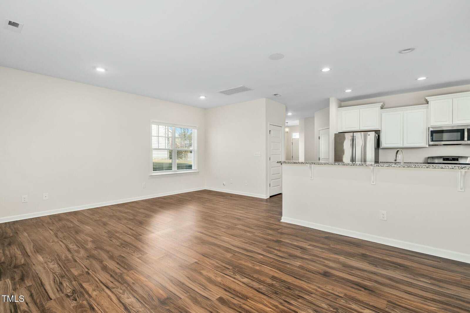 Open-concept living space with wood-style flooring, neutral walls, a window, and a kitchen area featuring white cabinets, stainless steel appliances, and a granite breakfast bar countertop.