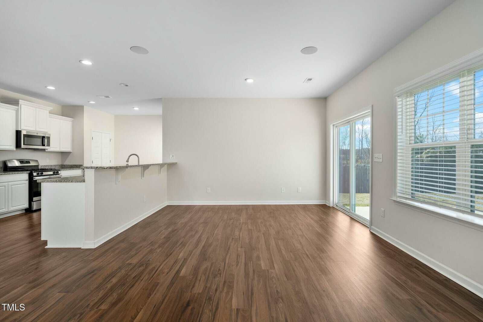 Bright, open-concept living space with wood flooring, white walls, large windows, and sliding glass doors leading to a backyard. The kitchen features white cabinets, granite countertops, and stainless steel appliances.