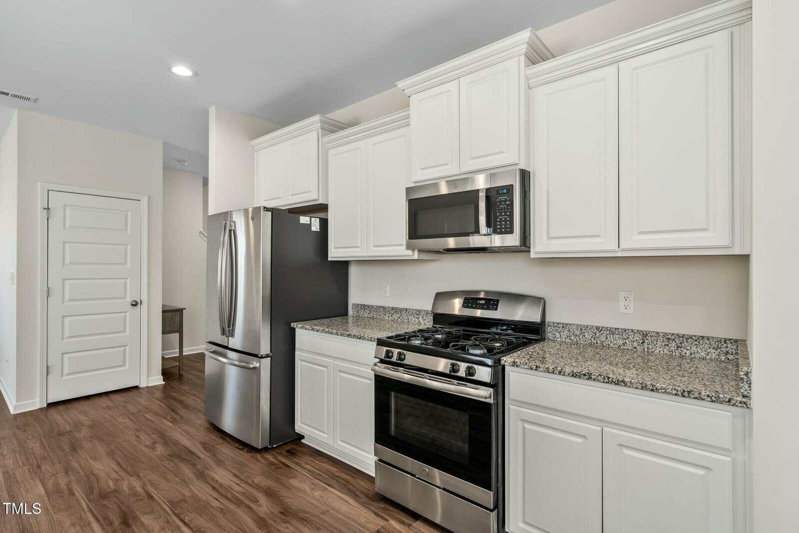Modern kitchen with white cabinets, stainless steel appliances including a refrigerator, microwave, and gas stove, granite countertops, and wood flooring. A white door and desk are visible in the background.