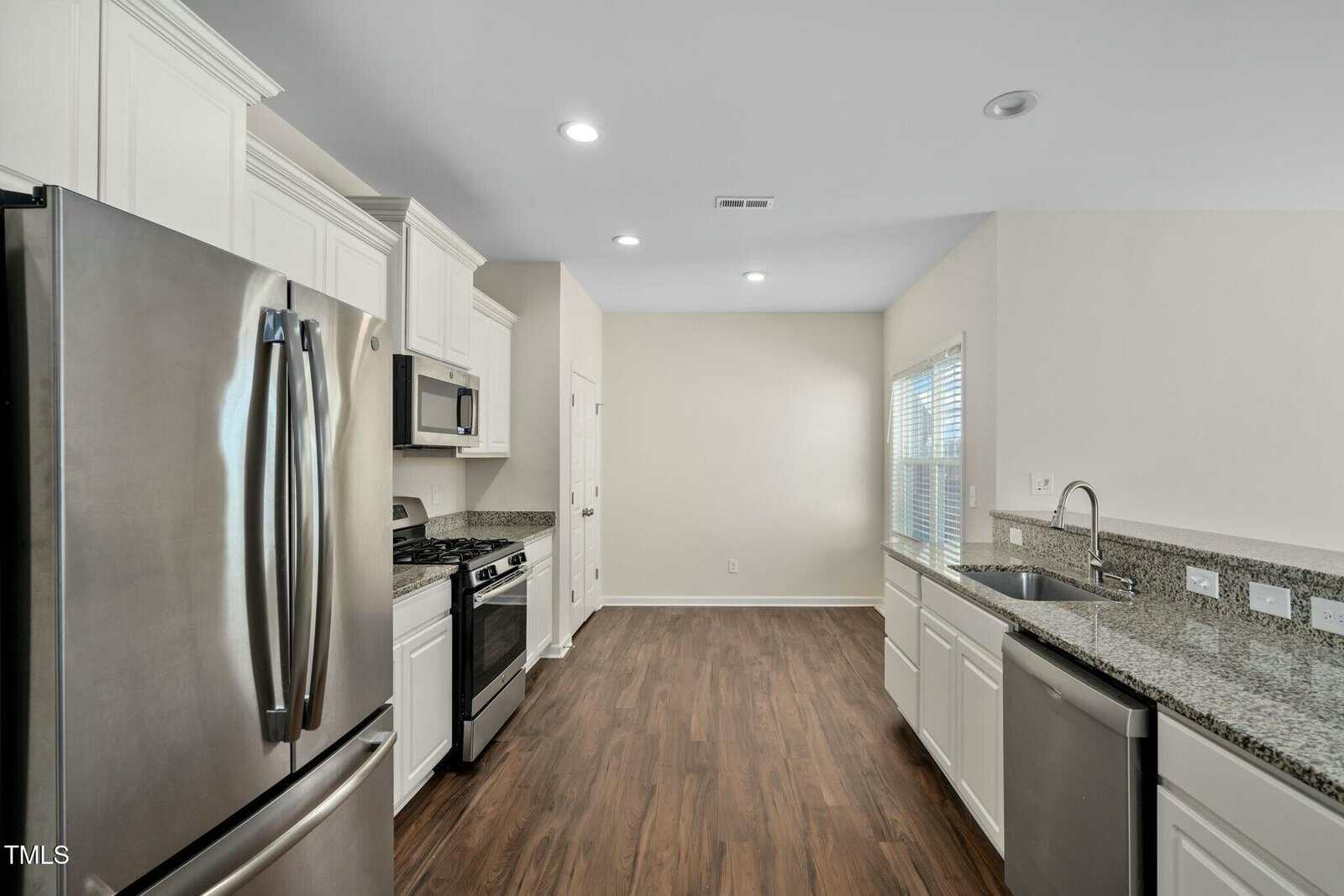 Modern kitchen with stainless steel appliances, white cabinets, granite countertops, and wood-style flooring. A window lets in natural light, and recessed lighting is installed in the ceiling.