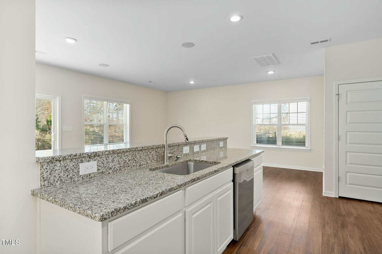 Modern kitchen with granite countertops, stainless steel sink, dishwasher, white cabinets, and wood-style flooring. Large windows provide natural light. Neutral walls and recessed lighting complete the space.