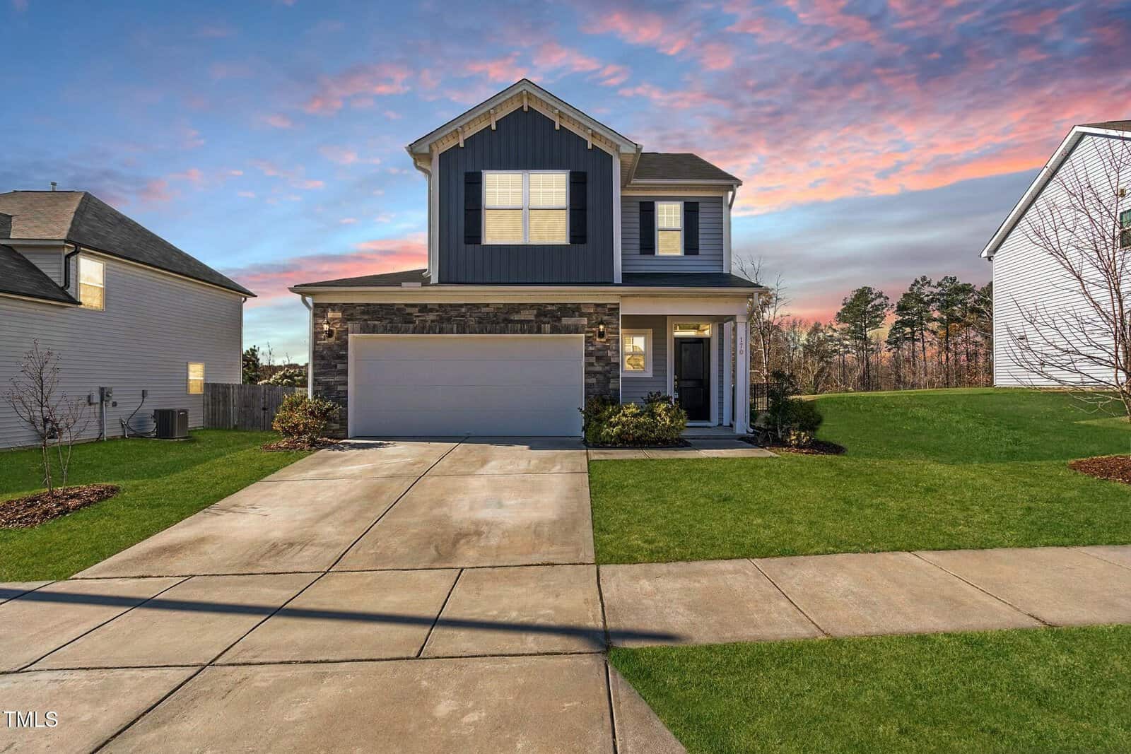 A two-story modern house with blue and gray siding, a double garage, and a stone accent facade. It sits on a well-manicured lawn at sunset, with a pink and blue sky in the background.