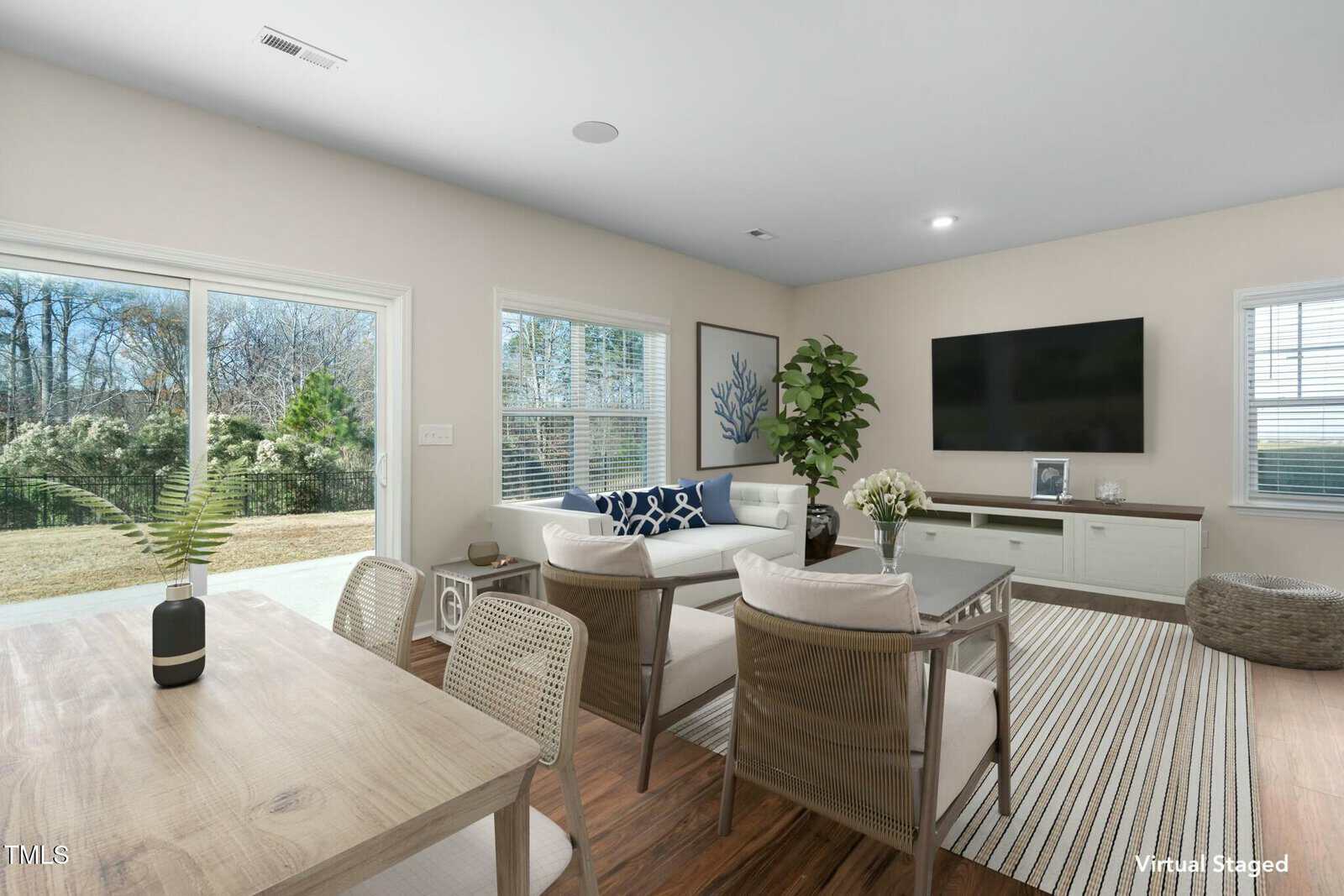 A modern living room with large windows, a wall-mounted TV, a white sofa, armchairs, a striped rug, indoor plants, and a dining table in the foreground. Sliding glass doors open to a grassy backyard.