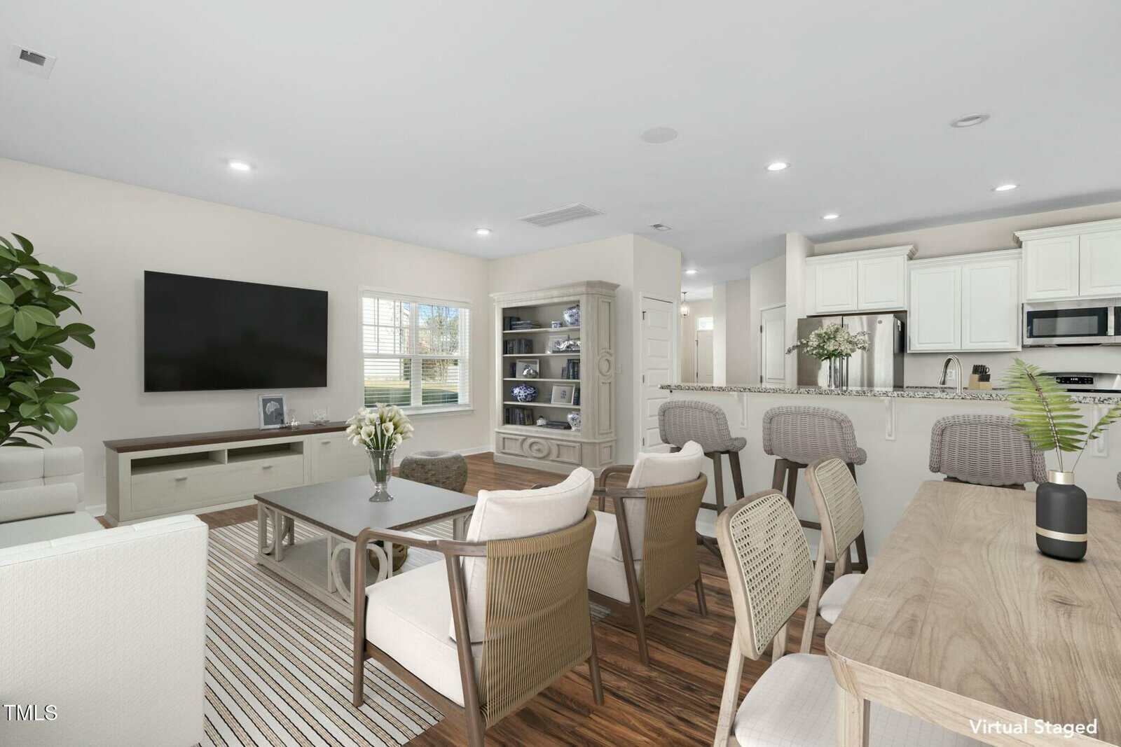 A bright, open-plan living and dining area with wood flooring, neutral furniture, a large wall-mounted TV, and a kitchen with white cabinets; sunlight streams in through a window. The image is virtually staged.