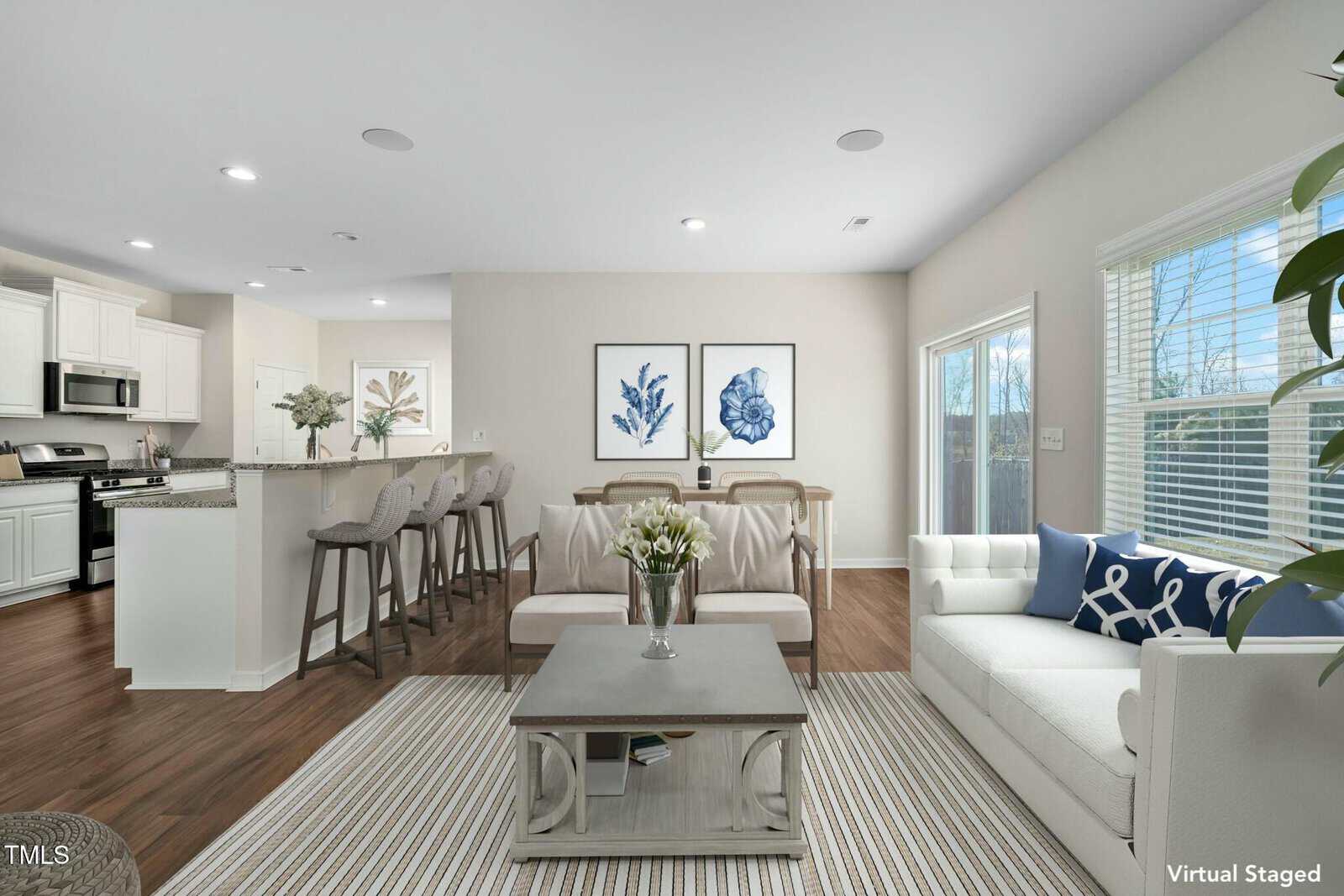 A modern, open-concept living room and kitchen with white cabinets, stainless steel appliances, a breakfast bar with stools, a white sofa, armchairs, a coffee table, floral art, and large windows letting in natural light.