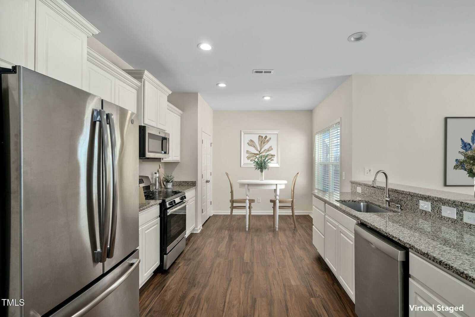 Modern kitchen with stainless steel appliances, white cabinets, granite countertops, and wood floors. A small dining table with two chairs is by a window, and framed artwork decorates the walls.