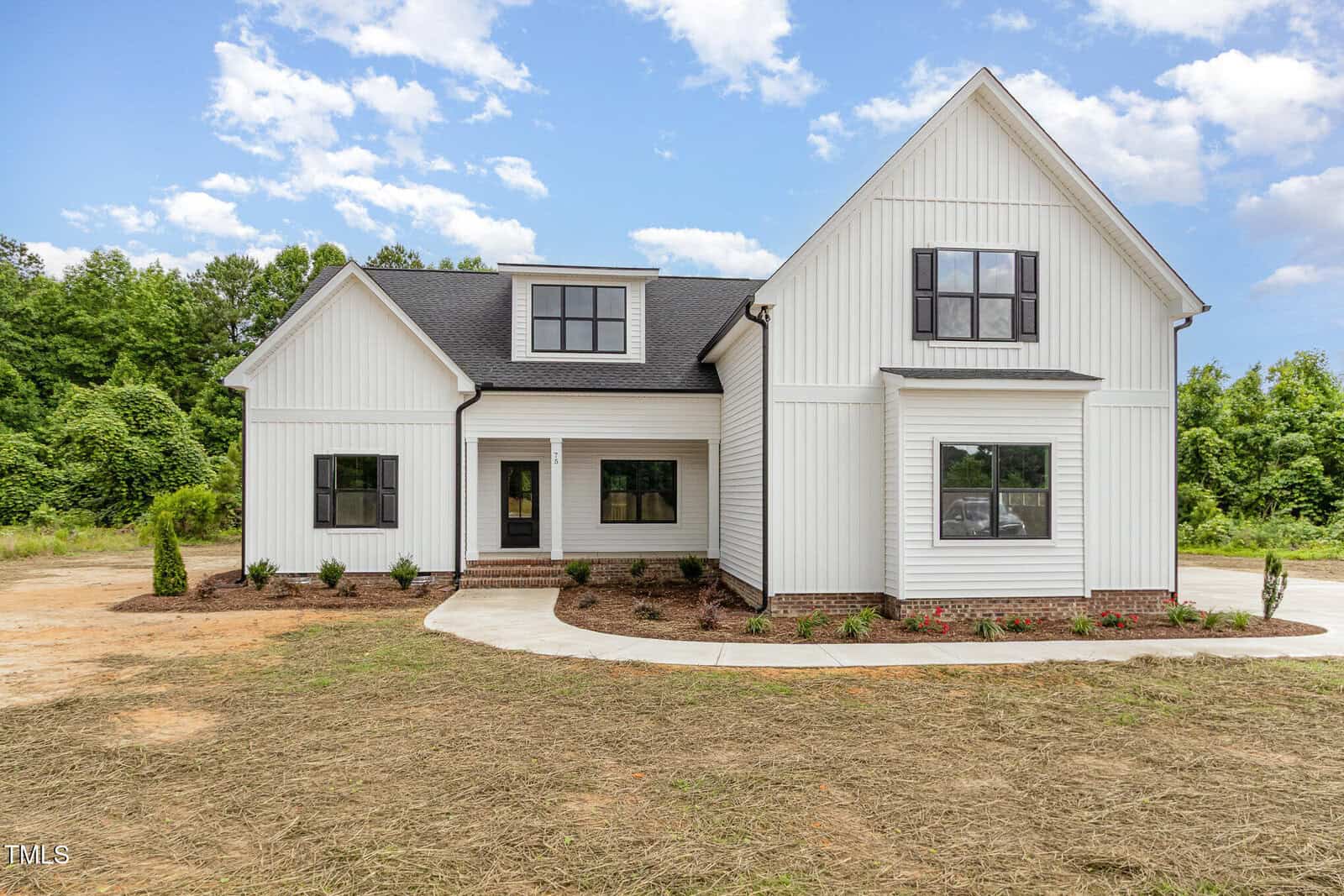 A modern white two-story house with black-framed windows, a front porch, and a landscaped yard with newly planted shrubs. The house sits on a lot with brown mulch and grass in front and trees in the background.