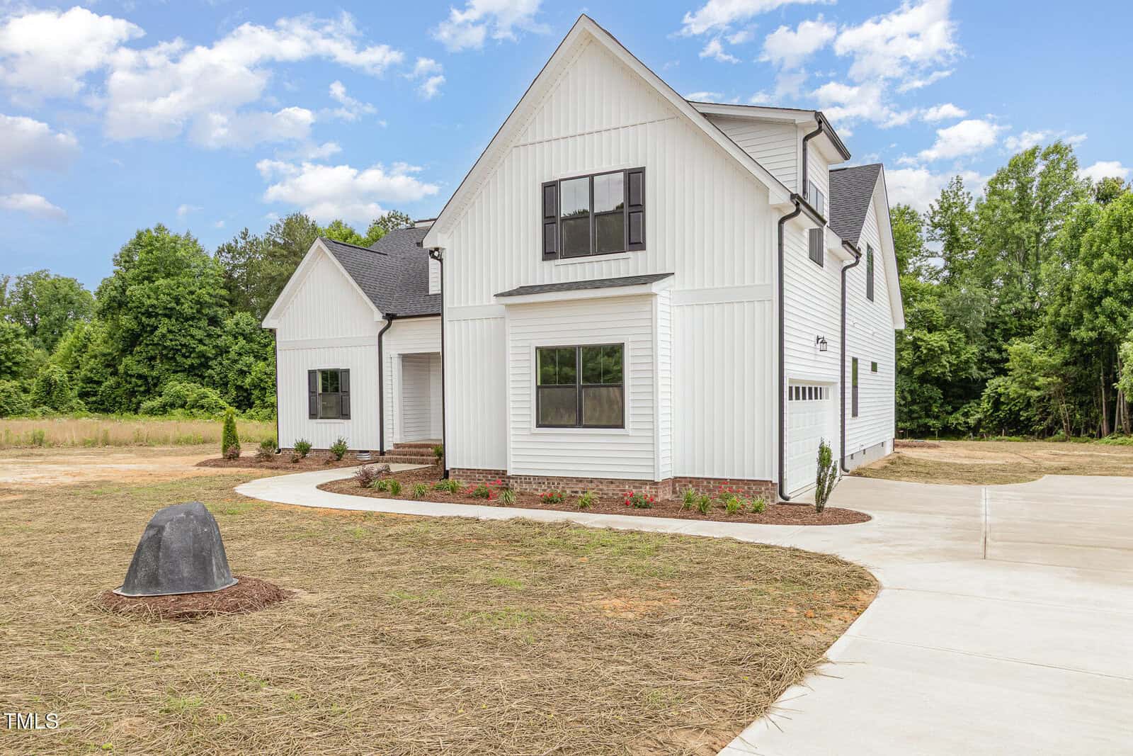 A modern two-story white house with black trim, a small covered porch, and landscaped shrubs in front. The driveway curves past the house, and the yard features straw-covered ground with a single rock and some trees in the background.