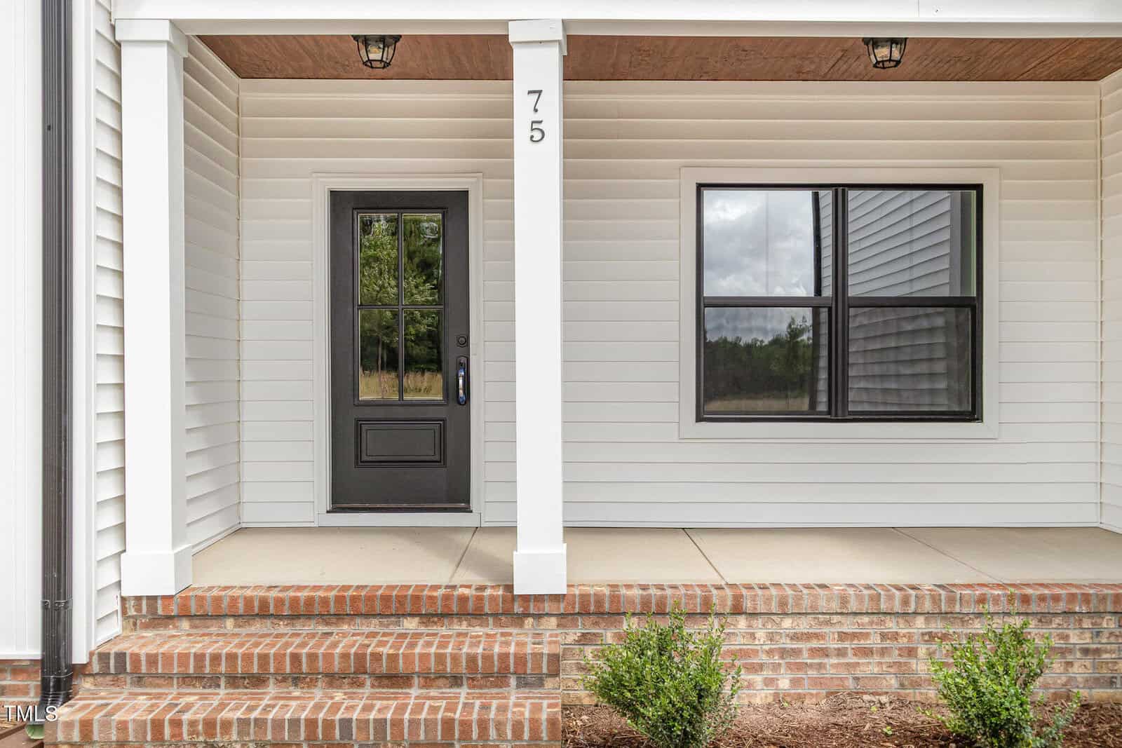 A small front porch with a black door, black-framed window, white siding, brick steps, and two ceiling lights. The porch has a white support column labeled with the number 75. Shrubs are planted in front.