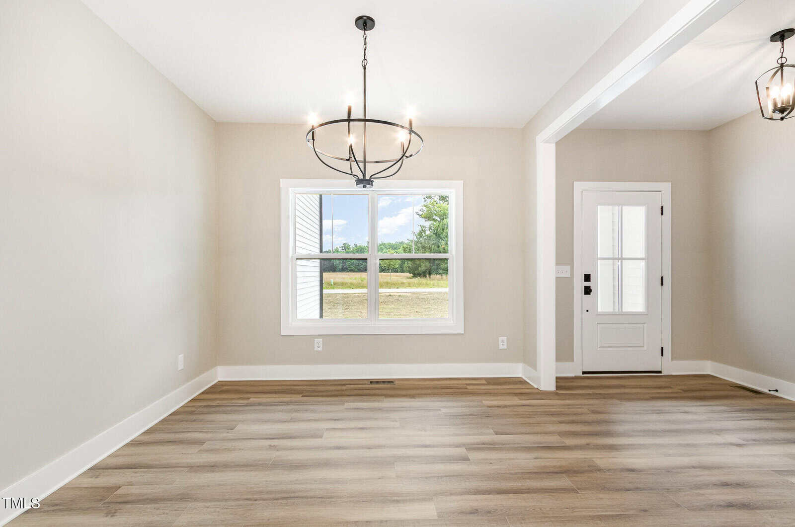 A bright, empty room with light wood flooring, beige walls, a large window showing green trees outside, a black chandelier, and a white door with glass panels and a black handle.