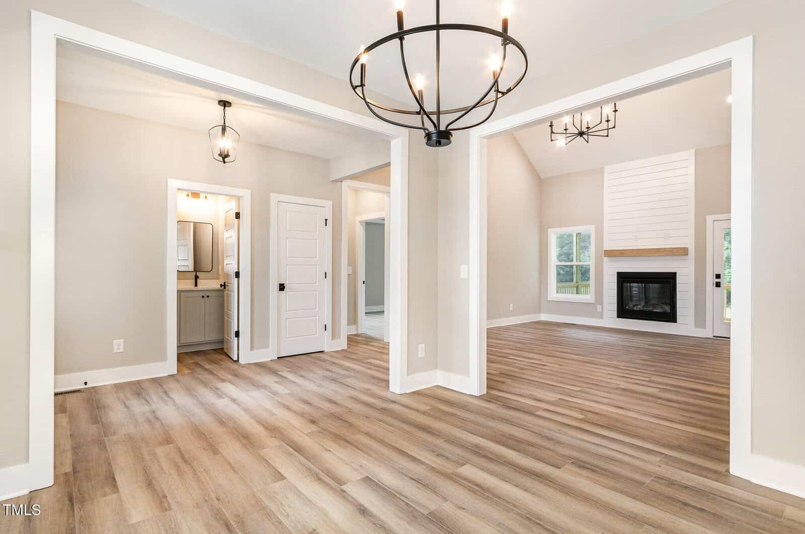Spacious modern home interior with light wood floors, neutral walls, two chandeliers, open living area with fireplace, and multiple white doors leading to other rooms. Large windows provide natural light.