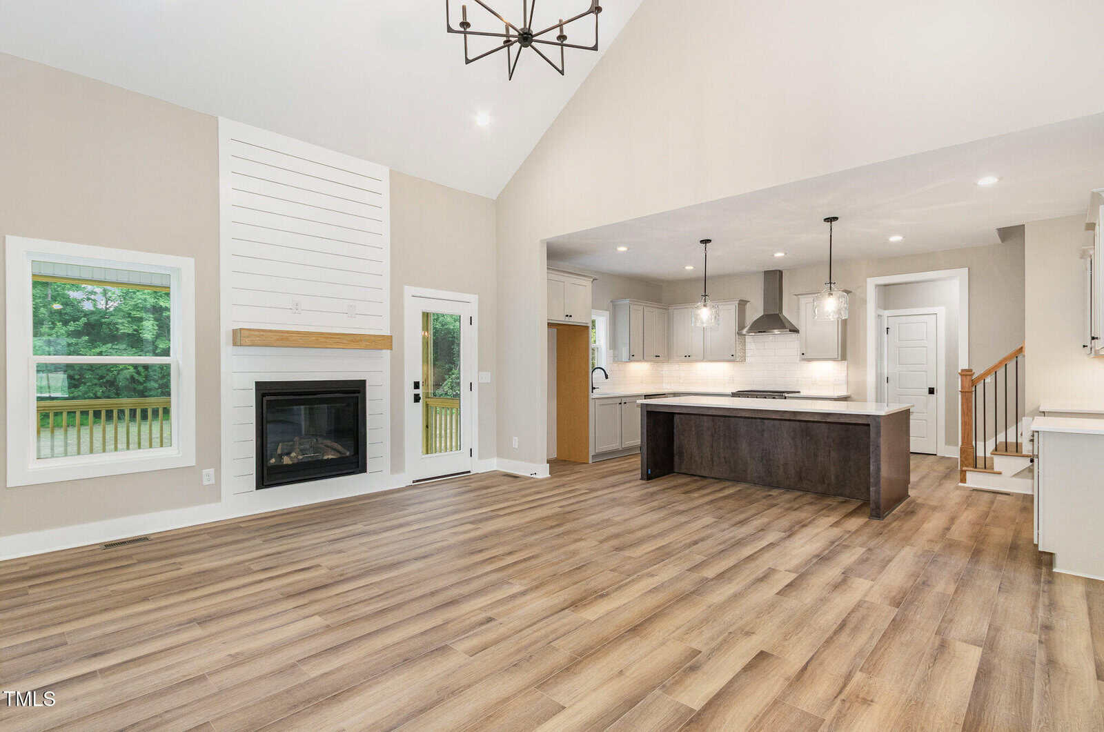 Modern open-concept living area with wood flooring, fireplace, large windows, and a kitchen featuring an island, pendant lights, white cabinets, and stainless appliances. Stairs lead up on the right side.