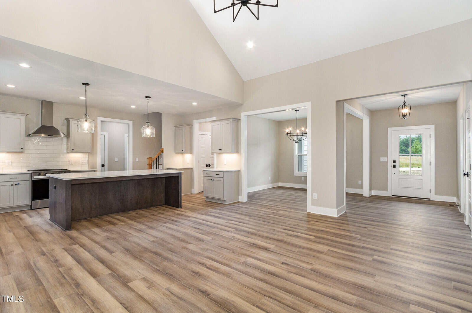 Open-concept kitchen and living area with wood floors, neutral walls, pendant lighting, island, white cabinets, and large windows letting in natural light. Dining area and entryway visible in the background.
