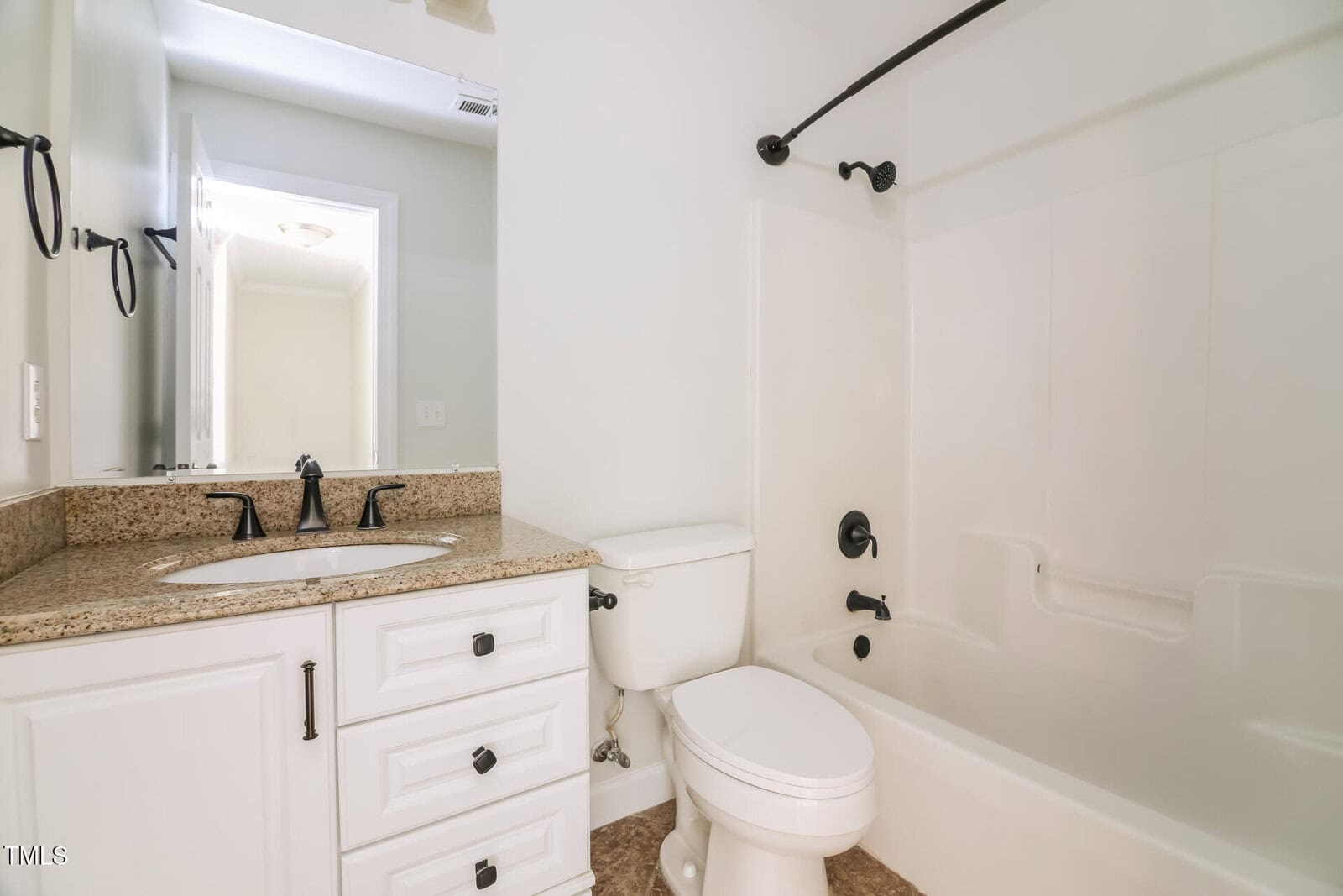 A bathroom with a white bathtub and shower, black fixtures, and a beige countertop with a sink. The vanity has white drawers, and a toilet is adjacent. A large mirror hangs above the sink, and the walls are painted light gray.