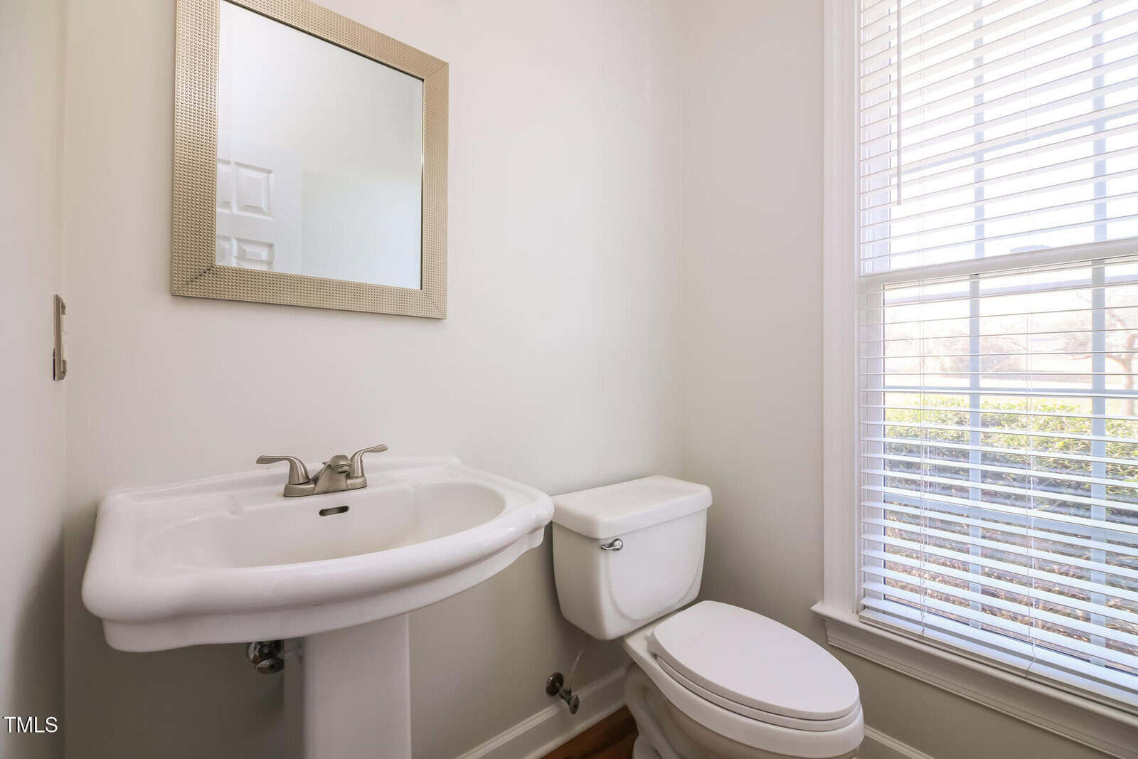 Small bathroom with a white pedestal sink and a toilet. A rectangular mirror with a light frame hangs above the sink. A window with white blinds is on the right, allowing natural light to enter the space. Walls are painted white.