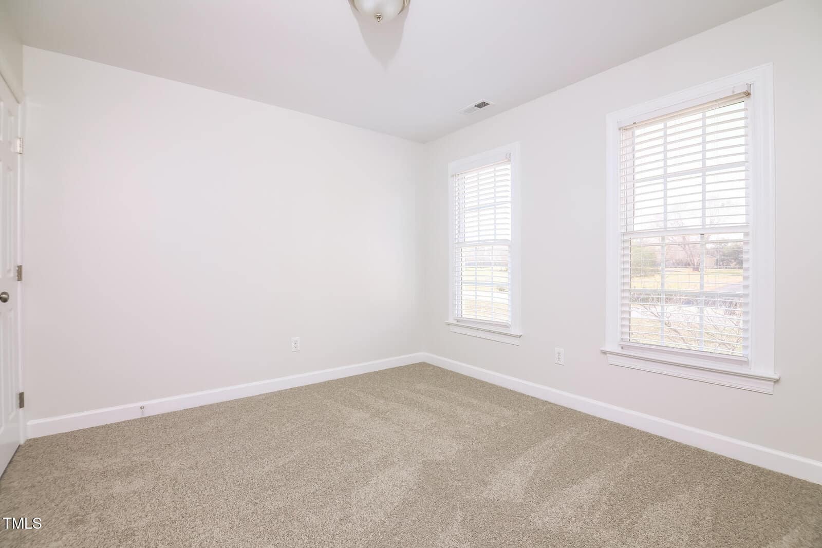 A small, unfurnished room with white walls and a beige carpet. Two windows with white blinds let in natural light. An overhead light fixture is on the ceiling, and part of a white door is visible on the left.