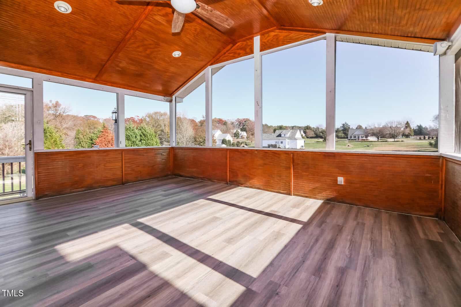 An empty sunroom with wooden ceiling and floor, featuring a ceiling fan. The windows provide a view of a suburban neighborhood with houses and trees in the distance under a clear sky.