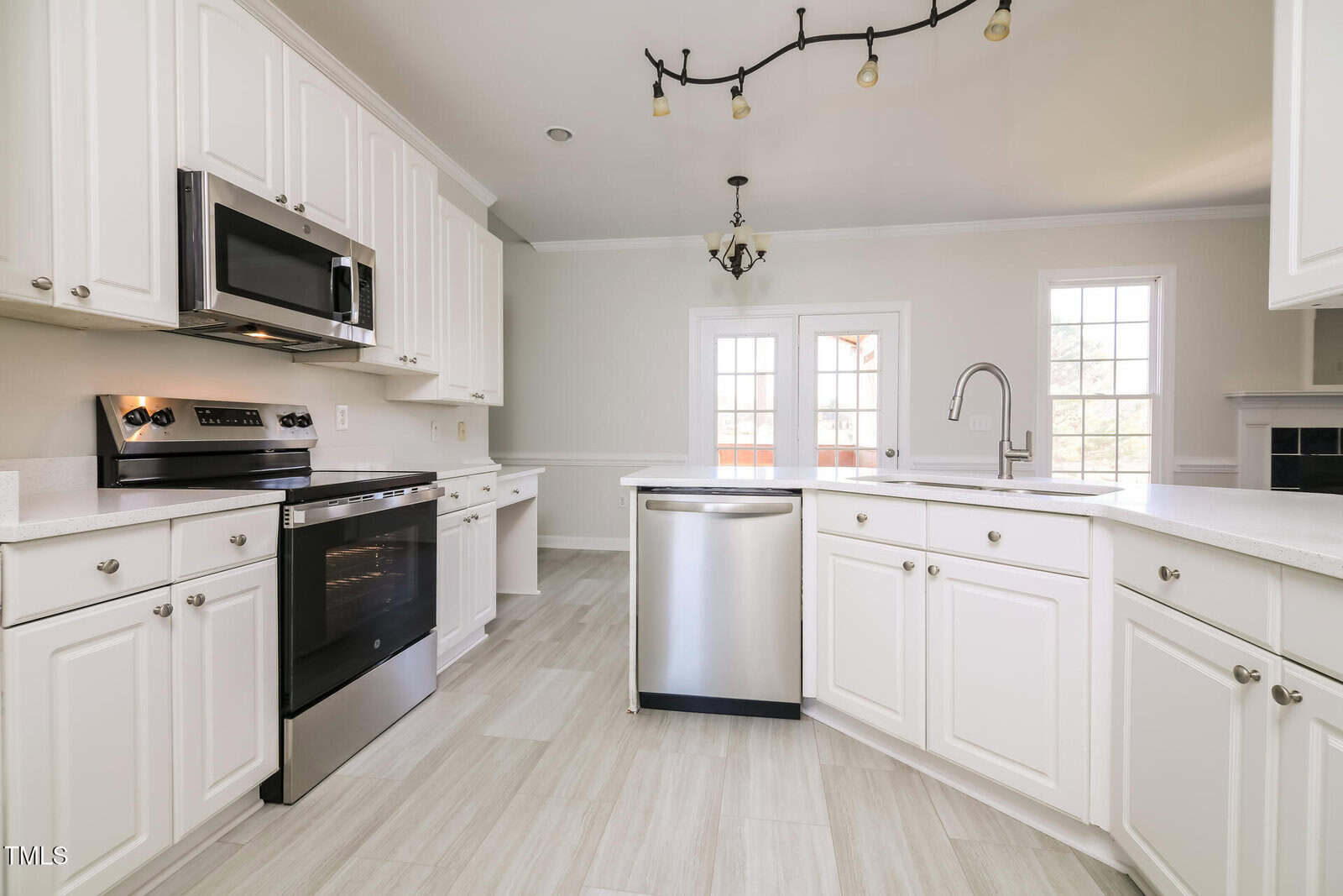 A bright kitchen with white cabinetry and tile flooring. It features a stainless steel oven, microwave, and dishwasher. A central island with a modern faucet is present. Two large windows and a glass door provide natural light.