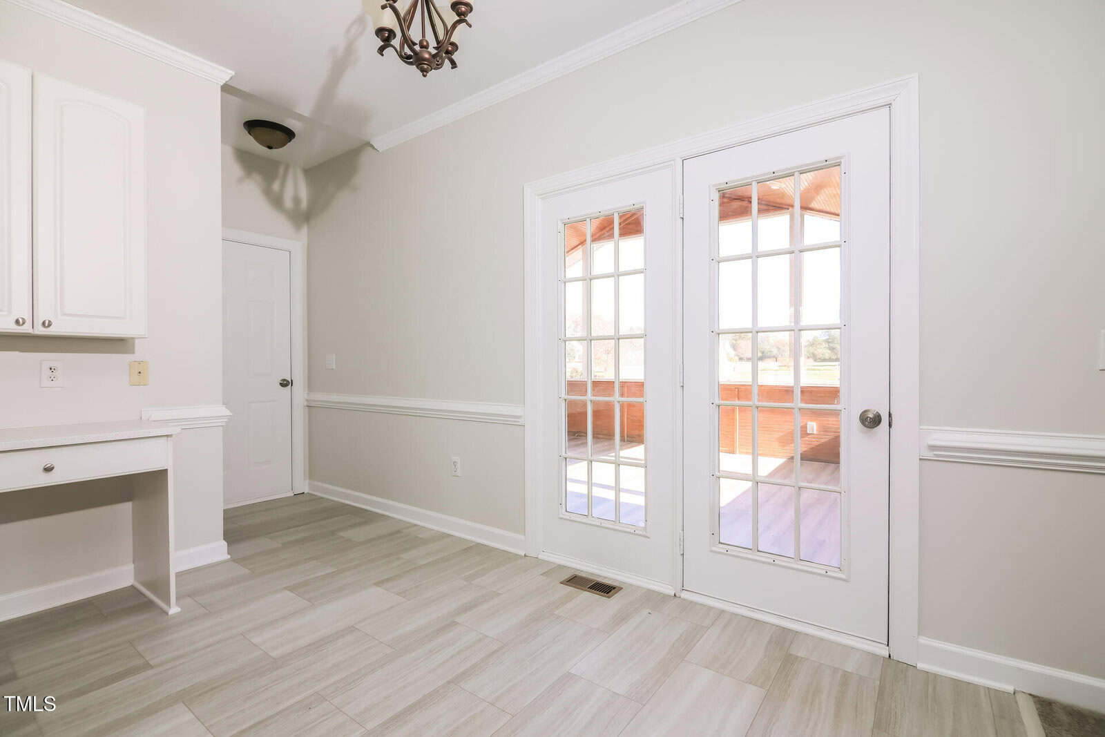 A bright room with light-colored tiled flooring, white walls, and a built-in desk to the left. French doors lead to a sunlit porch. A chandelier hangs from the ceiling, and a closed white door is on the left wall.
