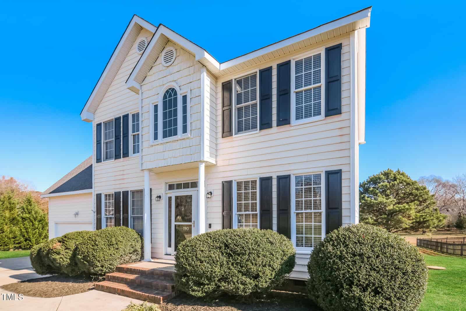 This charming two-story house at 190 Woodcrest Drive in Youngsville boasts a light-colored facade, dark shutters, and multiple windows. The inviting front entrance features a small porch with steps and shrubs on either side under the clear blue sky.