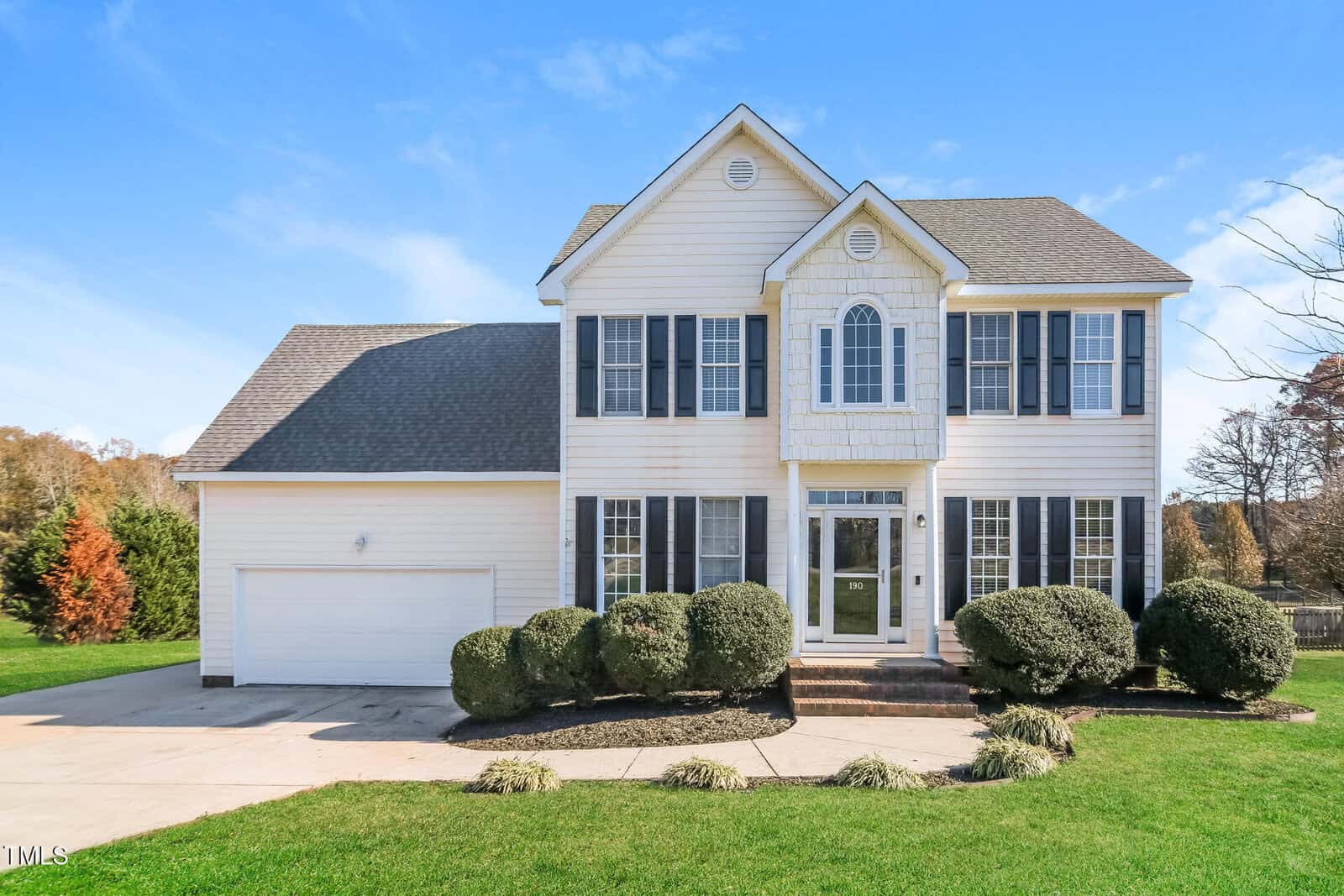 A two-story suburban house with light-colored siding and dark shutters. It features a gable roof, a two-car garage, and a manicured front lawn with shrubs. The entryway has a glass door with sidelights. A paved driveway leads to the garage.