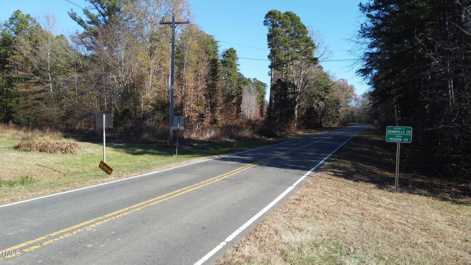 A rural road with double yellow lines runs through a wooded area. Leafless trees line the road under a clear blue sky. Two signs are visible: one points to "Oakville" and the other to "Person Co." Power lines run parallel to the road.