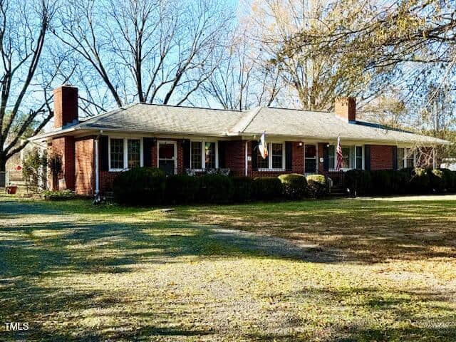 The single-story brick house at 812 Lewis Street, Oxford, boasts a gray roof, two chimneys, and a charming front porch adorned with two flags. The yard features lush grass and shrubs by the house, while trees with sparse foliage stand in the background.