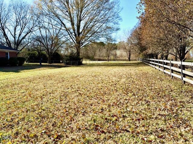 A grassy yard covered with fallen leaves extends toward a wooden fence on the right. Several large trees with bare branches are scattered across the property. The sky is clear and blue, and a red-brick house is partially visible on the left.