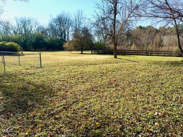A grassy field with sparse fallen leaves, bordered by a fence on three sides. Several trees with bare branches stand in the background. A chain-link fence encloses part of the field on the left side. The sky is clear and sunny.