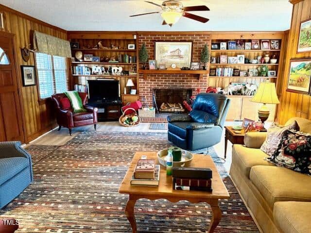 A cozy living room with wood-paneled walls and a brick fireplace. Shelves with books and decor flank the fireplace. The room features a striped rug, various armchairs, a couch with floral pillows, a ceiling fan, and a coffee table with books.