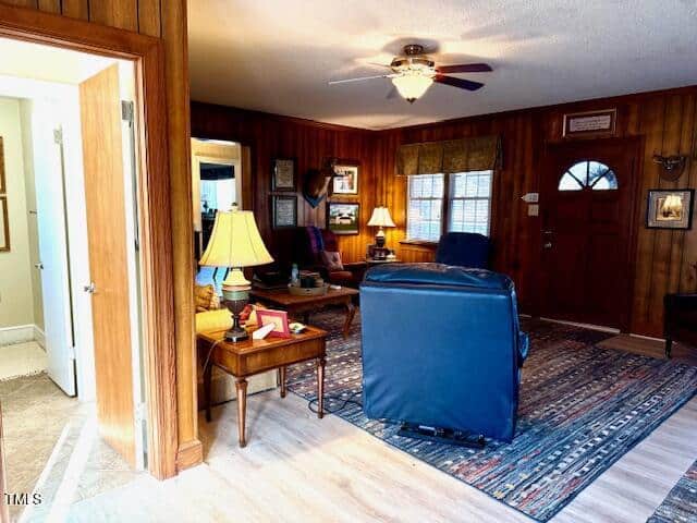 A cozy living room with wood-paneled walls features a tiled entryway, a blue recliner, and a plaid-patterned chair. A ceiling fan light is on, and natural light comes through a window. Decor includes lamps and wall art. A side table has books and a lamp.