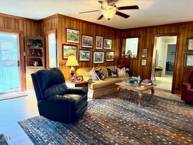 A cozy living room with wood-paneled walls features a ceiling fan and a brown sofa adorned with decorative cushions. A black recliner faces a wooden coffee table. Various framed pictures hang on the walls, and a colorful rug covers the floor.