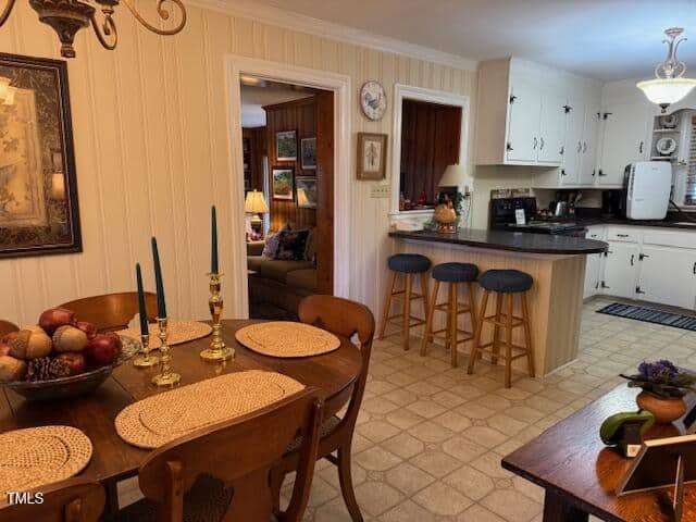 A cozy kitchen and dining area with beige walls and a tiled floor. The dining table has woven placemats, a bowl of fruit, and two green candles. The kitchen has white cabinets, a dark countertop, and three barstools. A living room is visible through a doorway.