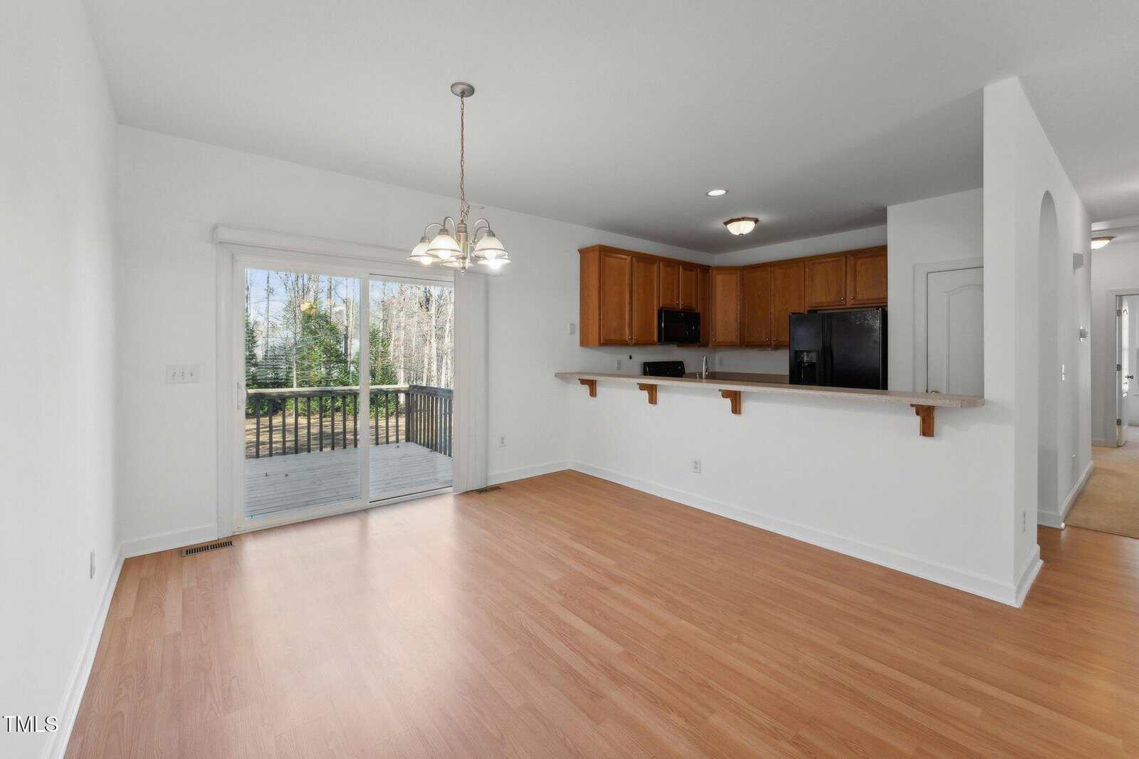 Bright dining area with wood floors, hanging light fixture, sliding glass doors opening to a deck, and an open kitchen with wooden cabinets and a breakfast bar. White walls and natural light fill the space.