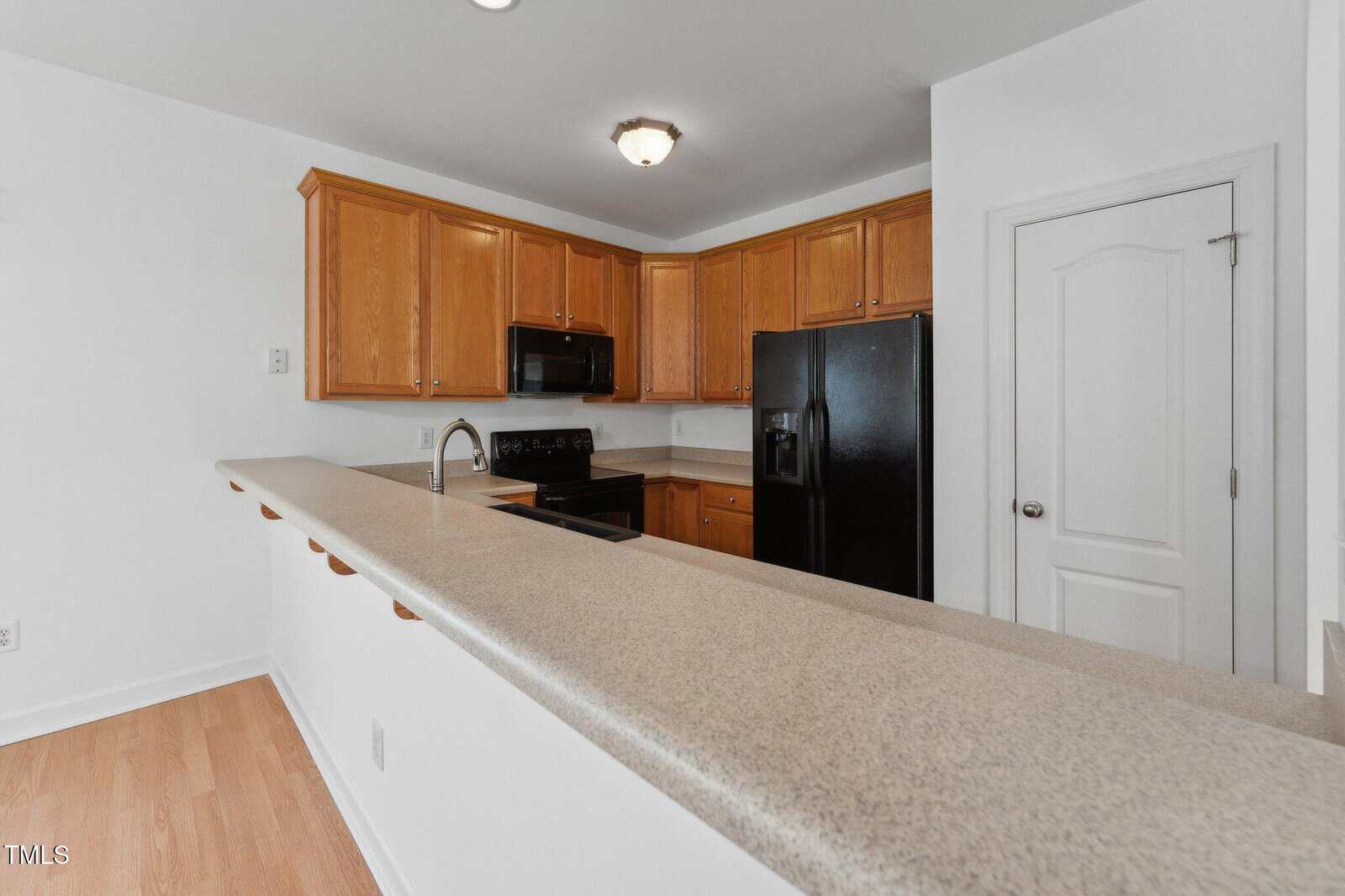 A kitchen with light wood cabinets, black appliances including a stove and refrigerator, a long beige countertop with a raised bar, and white walls with a closed white door.